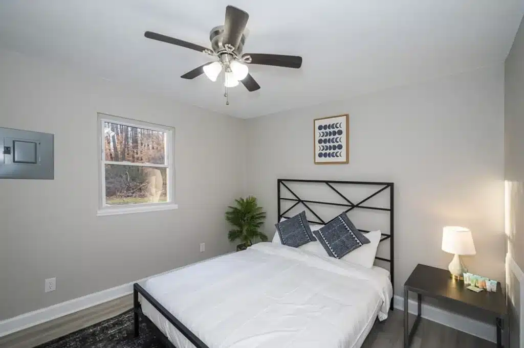 Modern bedroom with a double bed, white bedding, and two pillows. A nightstand with a lamp is beside the bed. Wall art hangs above, and a ceiling fan is on the white ceiling.