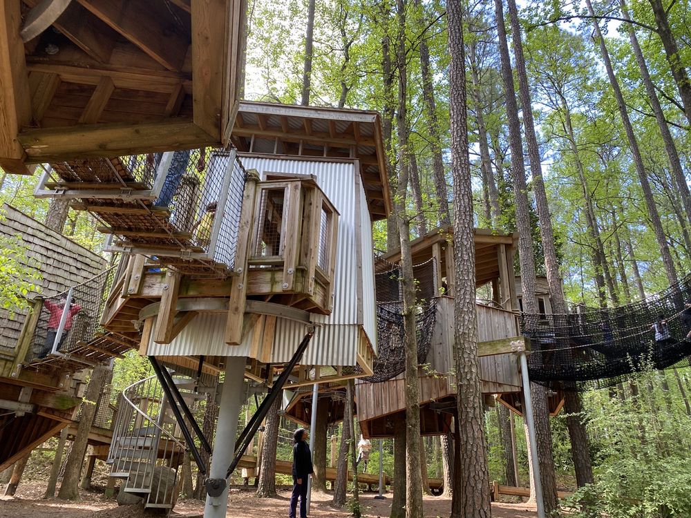Multiple wooden treehouses connected by rope bridges are elevated among tall trees in a forest, with a spiral staircase and people walking below.