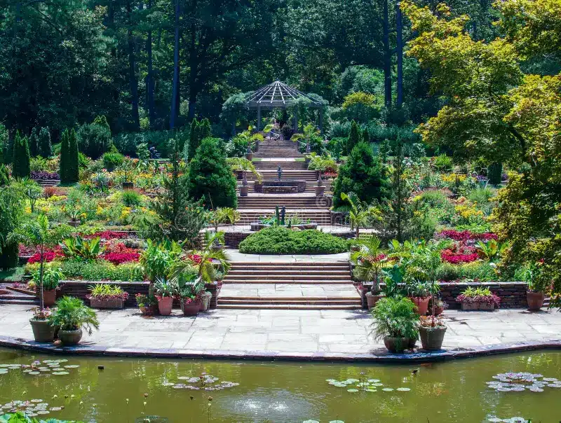 A landscaped garden with stone steps, vibrant flower beds, potted plants, and a pond with lily pads in the foreground. A gazebo structure is visible at the top of the stairs.